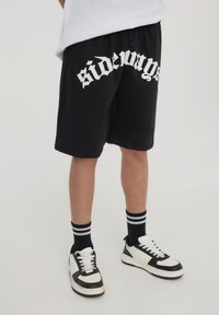 Pantaloni corti neri in cotone con stampa grafica bianca "di lato", cintura elastica, abbinati a sneaker bianche e nere e calze a righe nere.