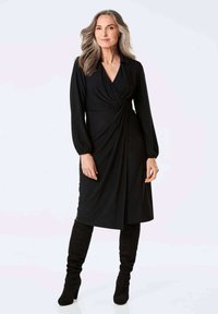 Schwarzes Wickelkleid mit langen Ärmeln, gerafftem Taillendetail und glatter Textur, kombiniert mit knielangen schwarzen Stiefeln mit Absatz.