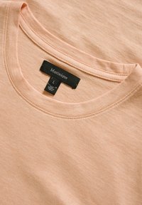 T-shirt de couleur pêche clair avec un col rond, texture côtelée et coutures piquées. Présente une étiquette noire avec des informations sur la marque et la taille.