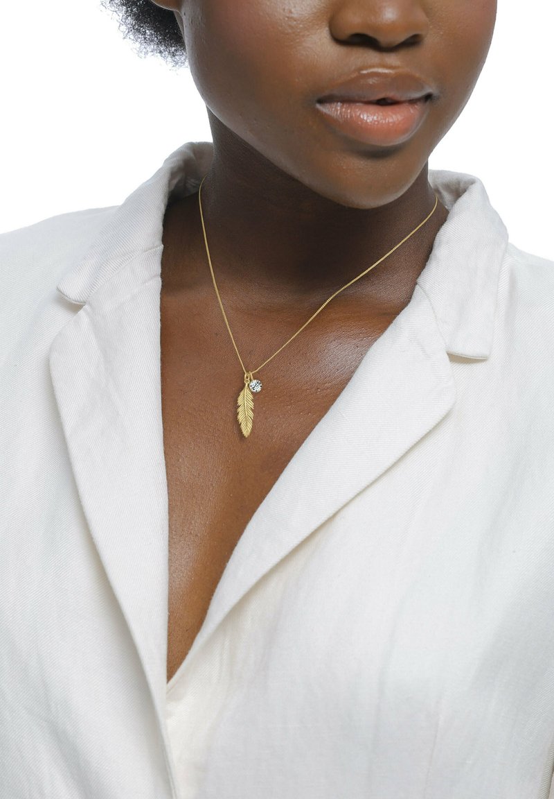 Goldenes Feder-Pendants mit einem kleinen klaren Edelstein an einer zarten Kette, getragen über einem weißen Hemd mit Kragen.