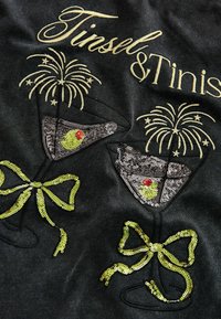 Czarna tkanina z haftowanym napisem „Tinsel & Tinis”, z cekinowymi szklankami martini, żółtymi akcentami i wzorami fajerwerków.