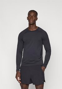 Melns sporta krekls ar garām raglāna piedurknēm, pieguļošs dizains, gluda tekstūra un smalka zīmola detaļa pie apakšmalas, kopā ar melniem šortiem.