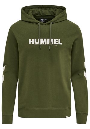 Kobieta w oliwkowo zielonej bluzie z kapturem Hummel i pasujących spodniach z białymi logo Chevron na rękawach oraz napisem "HUMMEL" na piersi.