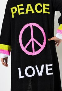 Schwarzer Strickpullover mit dem Wort "PEACE" in Gelb, einem pinkfarbenen Friedenssymbol und dem Wort "LOVE" in Weiß, und bunten, gestreiften Bündchen.