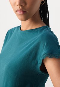 Teal bomull T-skjorte med rund hals og bølget blonderkant på korte ermer. Jevn tekstur, tettsittende fasong og subtil glans.