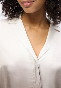 Κοντινό πλάνο ενός ατόμου με σγουρά μαλλιά που φοράει ανοιχτό μπεζ πουκάμισο με κουμπιά, χαμογελώντας απαλά με τα χείλη κλειστά.