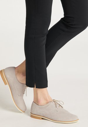 Light gray suede shoes with a rounded toe, flat wooden heel, and tonal laces, paired with black slim-fit pants featuring a slit hem.