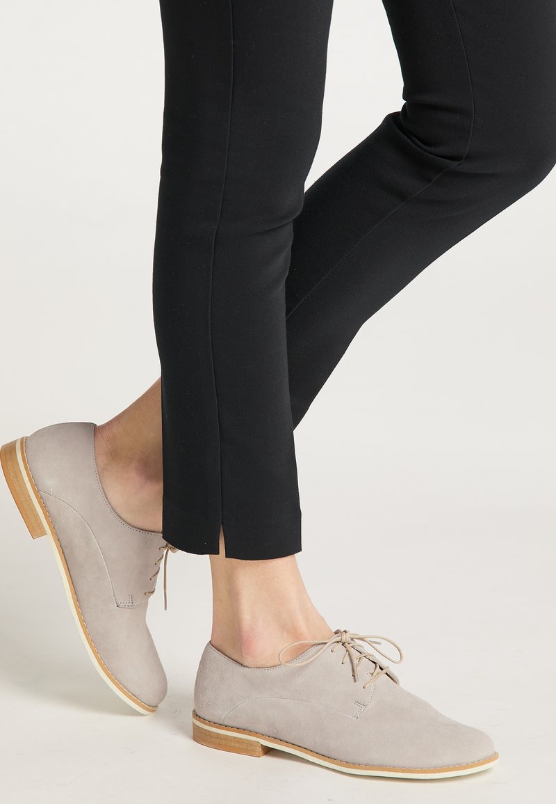 Lichtgrijze suède schoenen met een ronde neus, platte houten hak en ton-sur-ton veters, gecombineerd met zwarte slim-fit broek met een split aan de onderkant.