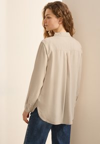 Camisa beige de manga larga con una textura suave, cuello y pliegue en la espalda. Combinada con pantalones de pana azules, mostrada desde atrás.