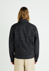 Black denim jacket with a classic collar, visible stitching, buttoned cuffs, and slightly textured fabric. Worn over light beige trousers.