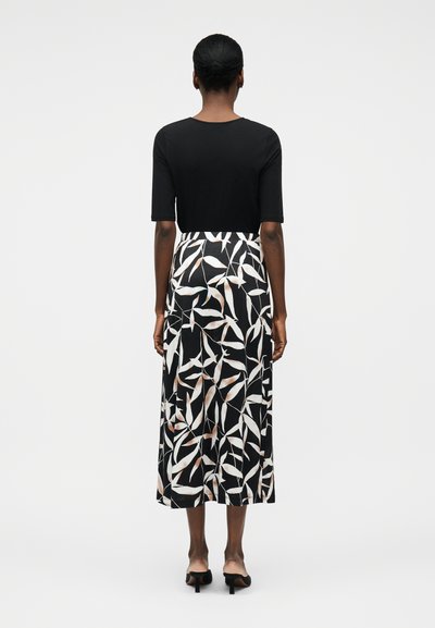 Γυναίκα με μαλλιά σε κότσο φορώντας μαύρο πάνω μέρος και μαύρη μεσαίου μήκους φούστα με λευκό και μπεζ σχέδιο φύλλων, στέκεται γυρισμένη προς τα πίσω σε λευκό φόντο.