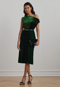 Grønn fløyelskjole med design på den ene skulderen, midtlang, innsnevret midje med et svart belte. Kombinert med en svart clutch og stroppede hæler.