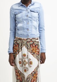 Light blue denim jacket with two front pockets, button closure, and long sleeves, worn over a patterned, flowy skirt featuring floral designs.