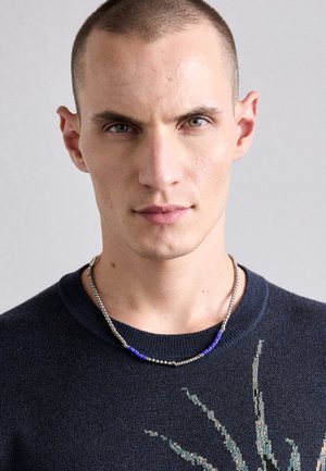 Jeune homme aux cheveux courts portant un pull foncé et un collier de perles argentées et bleues sur un fond clair uni.