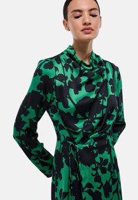 Robe fleurie verte et noire en tissu lisse. Présente un décolleté drapé et des manches longues avec un accent noué à la taille.