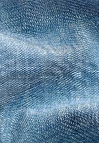 Denim tkanina u svijetloplavoj boji, s glatkom teksturom i suptilnim uzorkom tkanja. Nabranosti stvaraju slojeviti izgled.