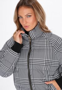 Giacca puffer in bianco e nero con motivo a scacchi houndstooth, colletto alto, chiusura con zip e polsini neri a coste. Texture liscia.