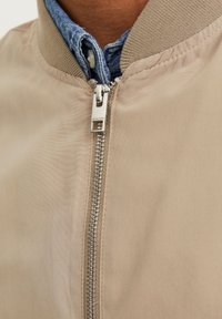 Beige bomberjacka med en slät texur, ribbat krage och silverdragkedja med en varumärkesetikett nära draget. Jeansskjorta under.