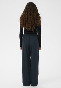 Femeie cu părul lung și ondulat, îmbrăcată cu un tricou negru cu mâneci lungi și pantaloni largi închiși la culoare, stând cu spatele într-un fundal alb.