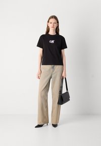 Zwarte T-shirt met korte mouwen met een logo, gecombineerd met brede beige jeans. Model houdt een zwarte handtas vast; zwarte puntige hakken maken de look compleet.