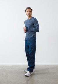 Modra pulover s motivom iz pletenine, z rebrastimi manšetami, v kombinaciji s temno modrimi kavbojkami in belimi športnimi čevlji. Ohlapen kroj, teksturiran material.