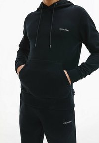 Sudadera negra con capucha de tela suave, con puños y dobladillo acanalados. Cuenta con un bolsillo frontal y un logo blanco de Calvin Klein en el pecho.