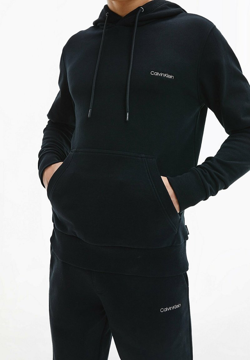 Sudadera negra con capucha de tela suave, con puños y dobladillo acanalados. Cuenta con un bolsillo frontal y un logo blanco de Calvin Klein en el pecho.