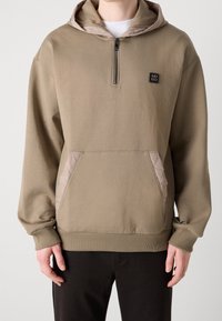 Khaki bluza z kapturem z półzamkiem i pikowanymi detalami na kieszeniach. Miękki materiał z prążkowanymi mankietami i obrębem. Naszywka z logo na piersi.