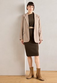 Beige blazer over a dark green ribbed dress with a belt. Tan suede ankle boots with a block heel. Smooth textures and tailored fit.