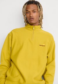 Geel fleece sweatshirt met een kwartrits, voorzien van een geborduurd logo in het bruin op de borst. Zachte textuur en ontspannen pasvorm.