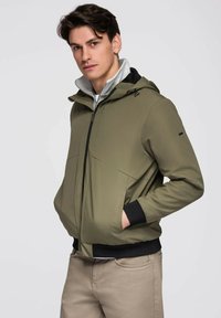 Młody mężczyzna w oliwkowozielonej kurtce z kapturem, z czarnymi mankietami i beżowymi spodniami, stojący z rękami w kieszeniach na jednolitym tle.