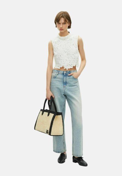 Femme portant un top court sans manches en dentelle florale blanche, un jean taille haute bleu clair, des chaussures noires, et tenant un sac à main en toile beige avec des bordures noires.