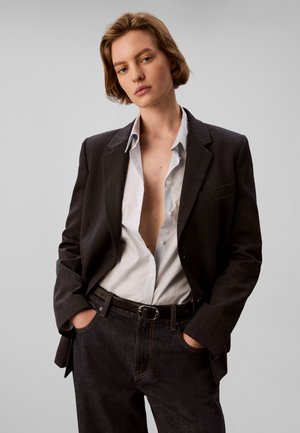 Jeune personne aux cheveux courts portant un blazer foncé sur une chemise blanche ouverte et un jean foncé, debout les mains dans les poches.