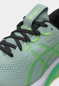 Κοντινή λήψη του παπουτσιού τρεξίματος ASICS GEL-Cumulus 28 σε πράσινο και μαύρο χρώμα, με πλέγμα στο πάνω μέρος, μαύρα κορδόνια και νεονκίτρινες λεπτομέρειες.