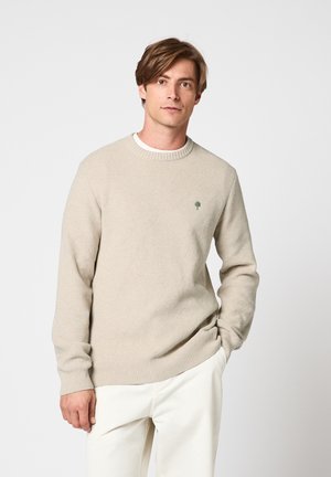 Jeune homme aux cheveux châtain clair portant un pull en maille beige et un pantalon blanc, debout avec une main dans la poche sur un fond uni.