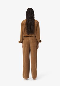 Camicia in velluto corduroy marrone e pantaloni a gamba larga, tessuto testurizzato, vista da dietro, capelli lunghi, cuciture visibili e due tasche posteriori sui pantaloni.