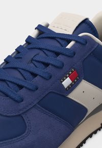 Baskets bleu et beige avec des lacets bleus, un tissu texturé et des panneaux en daim, arborant un petit logo Tommy Jeans sur le côté.