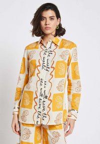 Lightweight button-up shirt in cream with yellow and brown abstract floral patterns and handwritten text; long sleeves and a classic collar.