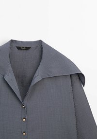 Chemise bleu marine et blanche à fines rayures, avec col large et boutons dorés, présentée sur fond blanc.