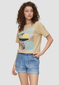 Beige Baumwoll-T-Shirt mit einem bunten Grafikdruck einer Strandlandschaft. Kombiniert mit hellen Denim-Shorts mit elastischem Bund und Seitentaschen.