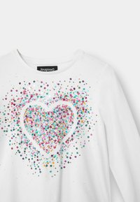 White long-sleeve shirt with a multicolored glitter heart design. Features vibrant pink, teal, and gold sequins scattered around the heart.