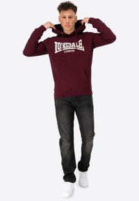 Bordeaux hoodie met een voorzak en een wit "Lonsdale London" logo. Gecombineerd met zwarte jeans en witte sneakers. Model past de capuchon aan.