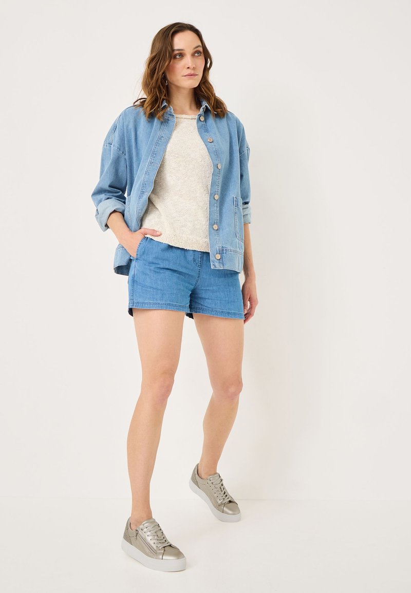 Giacca di denim di un blu chiaro, maglione di maglia beige, pantaloni corti blu e sneakers grigio metallico. L'outfit presenta uno stile casual e rilassato.