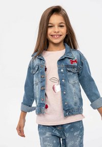 Jeans jakna ima klasičen kroj s poškrobnima žepoma na prsih in rdečimi češnjevimi aplikacijami. Nosimo jo preko svetlo roza srajce s sequinskimi detajli.