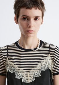 Gestreiftes T-Shirt in Schwarz und Creme mit einem durchsichtigen Spitzenoverlay und dekorativem Spitzenbesatz. Eine silberne Kettenhalskette ergänzt das Design.