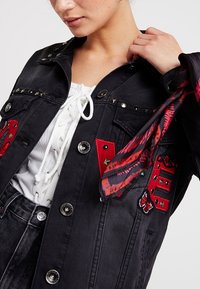 Czarna kurtka dżinsowa z czerwonymi naszywkami, metalowymi ćwiekami i wzorzystym szalikiem. Zapięcia na guziki oraz teksturowany wzór materiału.