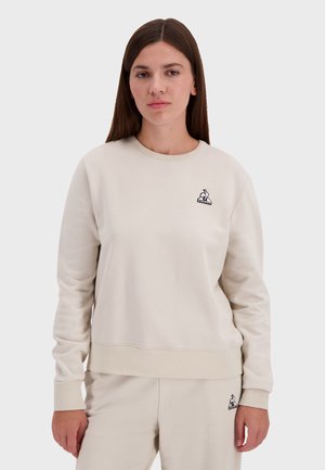 Beige crewneck sweatshirt van zacht materiaal, met een klein zwart konijnenlogo op de linkerkant van de borst en ribgebreide manchetten en zoom.