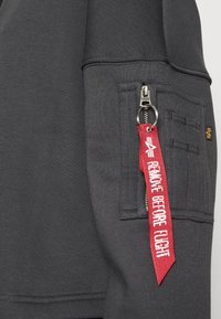 Ciemnoszary rękaw bluzy z kapturem posiada kieszeń na zamek błyskawiczny oraz czerwony naszywka z napisem "REMOVE BEFORE FLIGHT". Miękka faktura, ściągacze przy mankietach.