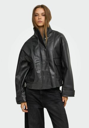 Jeune femme portant une veste en cuir noire oversize avec un col haut et un pantalon noir, debout devant un fond clair uni.