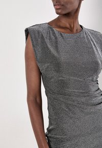 Vestido metálico plateado, sin mangas, con escote redondeado y textura fruncida. La tela suave y brillante se drapea elegantemente sobre el cuerpo.